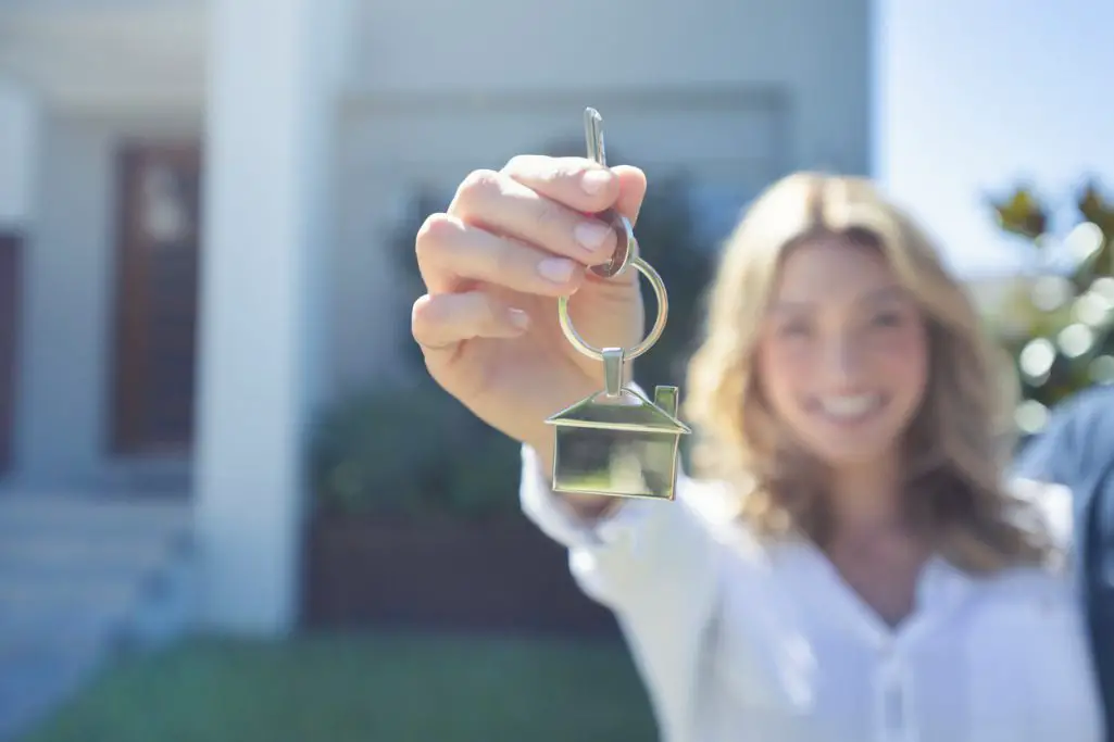 woman-smiling-and-holding-house-key-towards-camera-while-standing-in-front-of-house